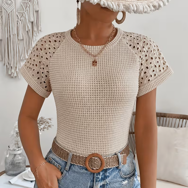 A woman wearing a beige short-sleeve knit top with eyelet shoulder details, paired with denim shorts and a woven belt, posed indoors with a wicker chair and macrame decor in the background.