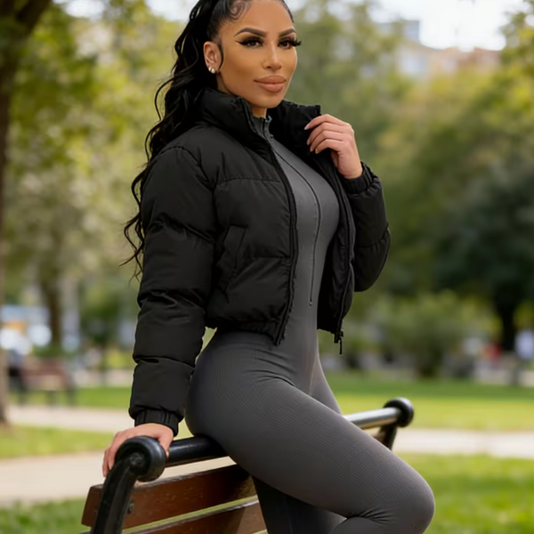 A woman posing on a park bench wearing a black cropped puffer jacket, a gray ribbed knit jumpsuit, and gray sneakers.